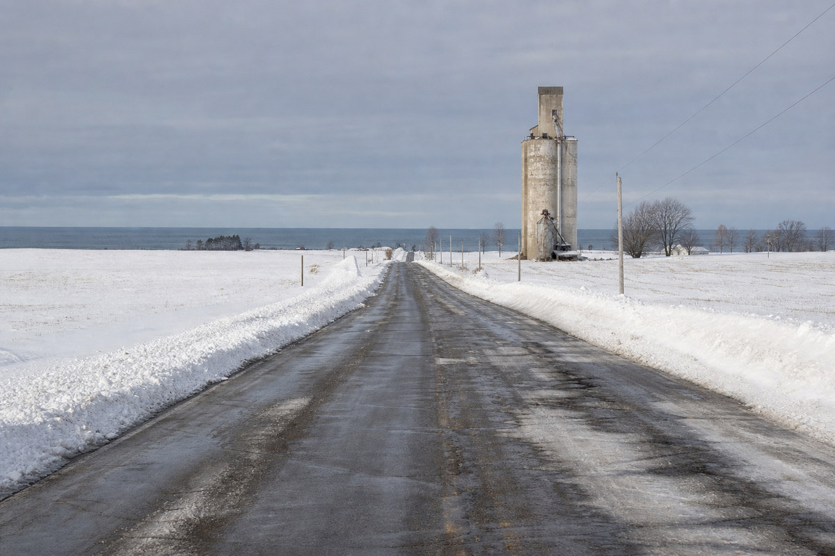 A wide flat Erie County road after a lake effect snowfall with plowed banks on both shoulders, a grain elevator and a low gray sky over Lake Erie